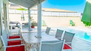 Una zona exterior coberta amb una piscina, cadires blanques, una taula amb estova floral i plantes en testos.