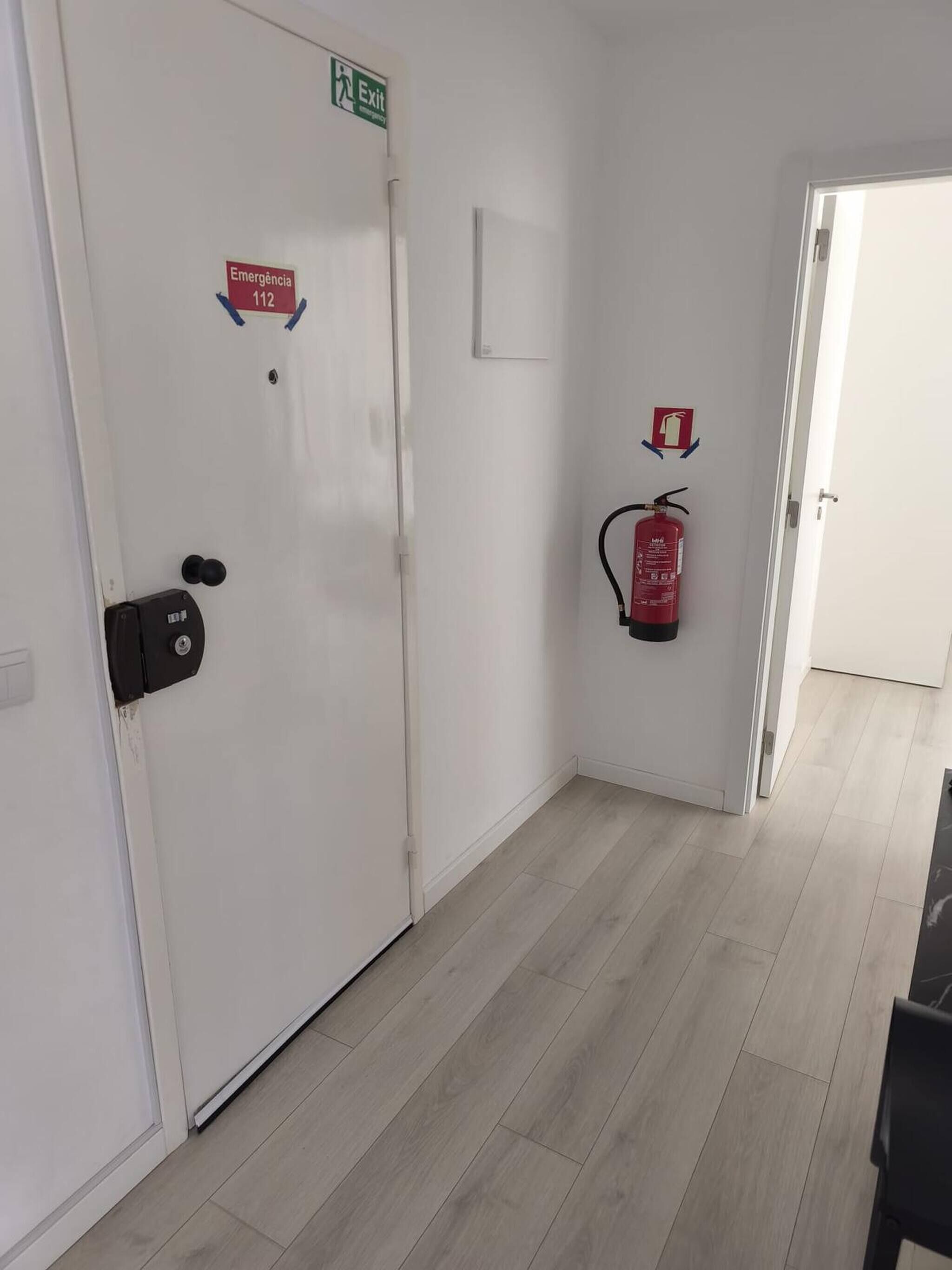 Un pasillo con una puerta blanca señalizada con “Emergência 112” y un letrero de salida, un extintor y una puerta que da a otra habitación.