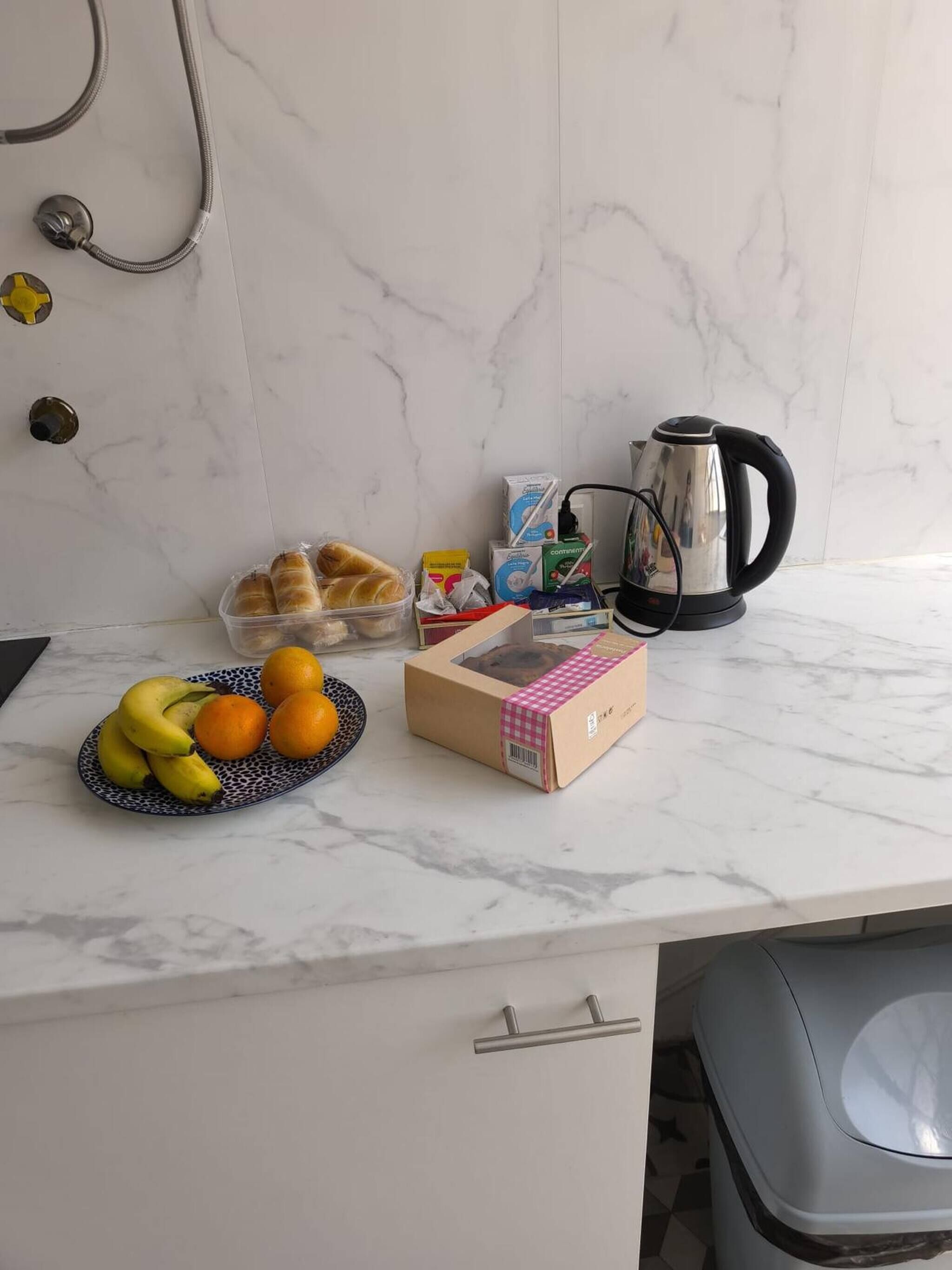 A kitchen counter with a marble backsplash, a kettle, a box of bread, a plate of fruit, and a trash bin.