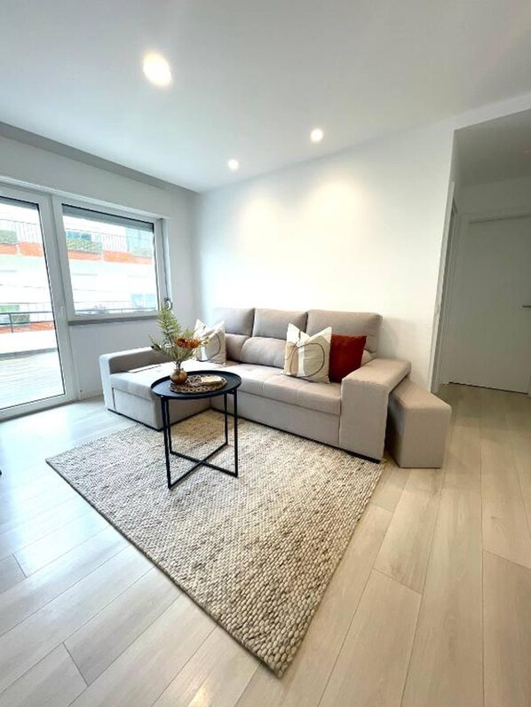Ein modernes Wohnzimmer mit einem grauen Sofa, einem kleinen runden Tisch und einem großen Fenster.
