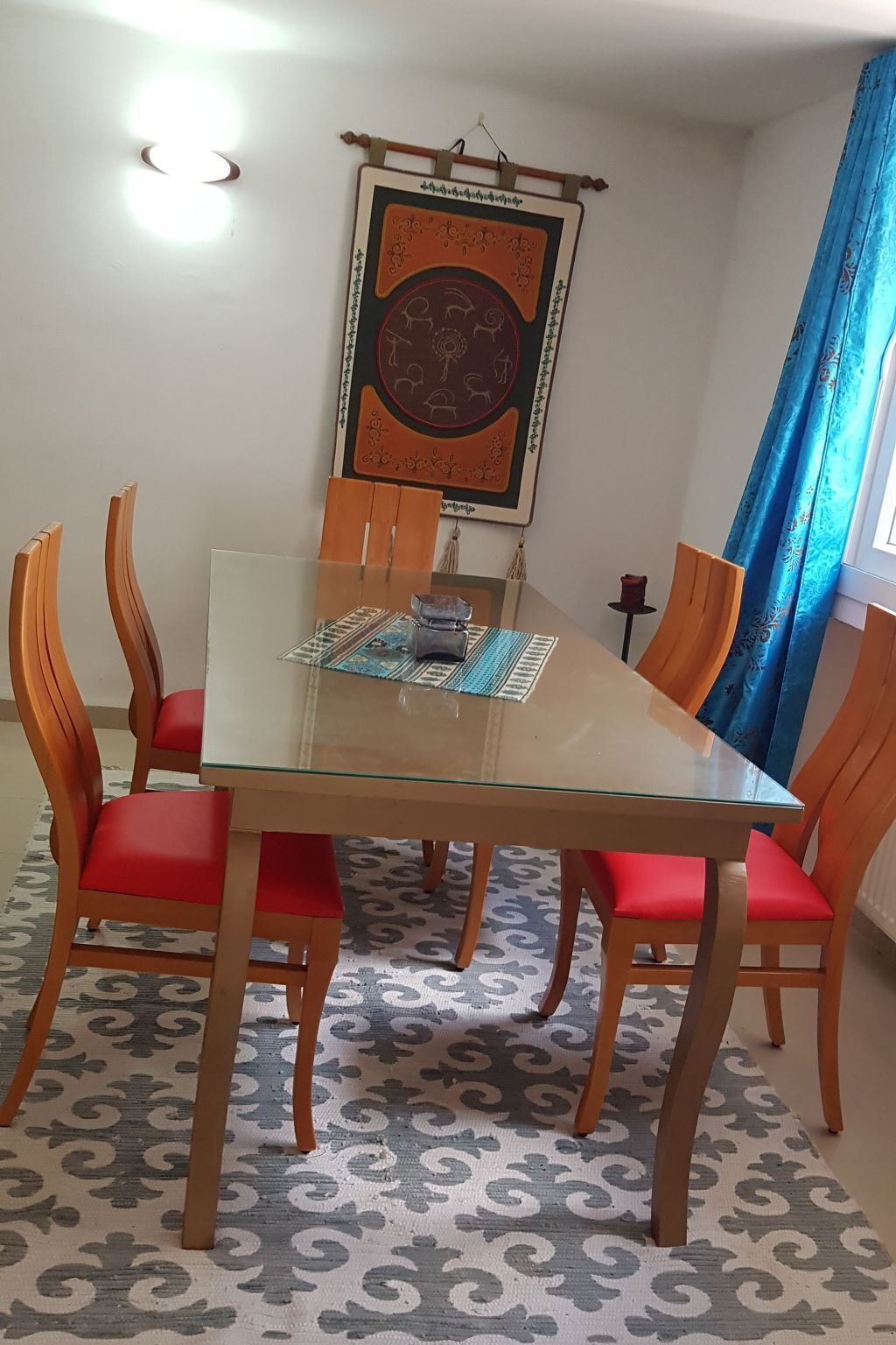 Un coin repas comprenant une table à plateau en verre, six chaises en bois avec des coussins rouges, un tapis à motifs et un élément décoratif fixé au mur.