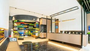 Un área de recepción moderna con una estantería colorida, un espacio de descanso con un banco y sillas, y un mostrador con pantalla digital.