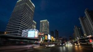 Un paisaje urbano nocturno con edificios altos, una calle concurrida y vehículos en movimiento, además de vallas publicitarias iluminadas.