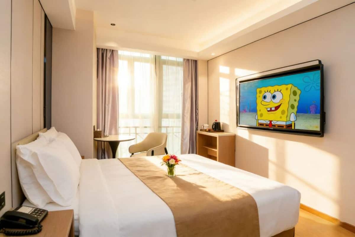 Une chambre d’hôtel avec un lit, un téléviseur diffusant l’émission « Bob l’éponge », une table de chevet avec un téléphone et une petite table sur laquelle est posé un vase de fleurs.