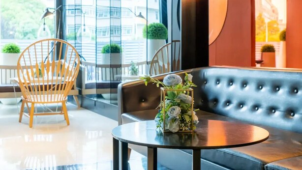 Un moderno vestíbulo de hotel con un sofá de cuero negro, una silla de madera, una mesa redonda con un centro de mesa floral y ventanas grandes con vistas a la ciudad.