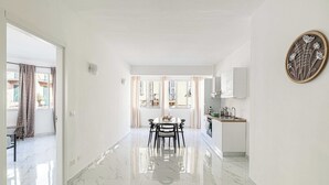 Una cuina moderna amb una illa central, una zona de menjador amb taula i cadires, i un passadís que condueix a un dormitori.
