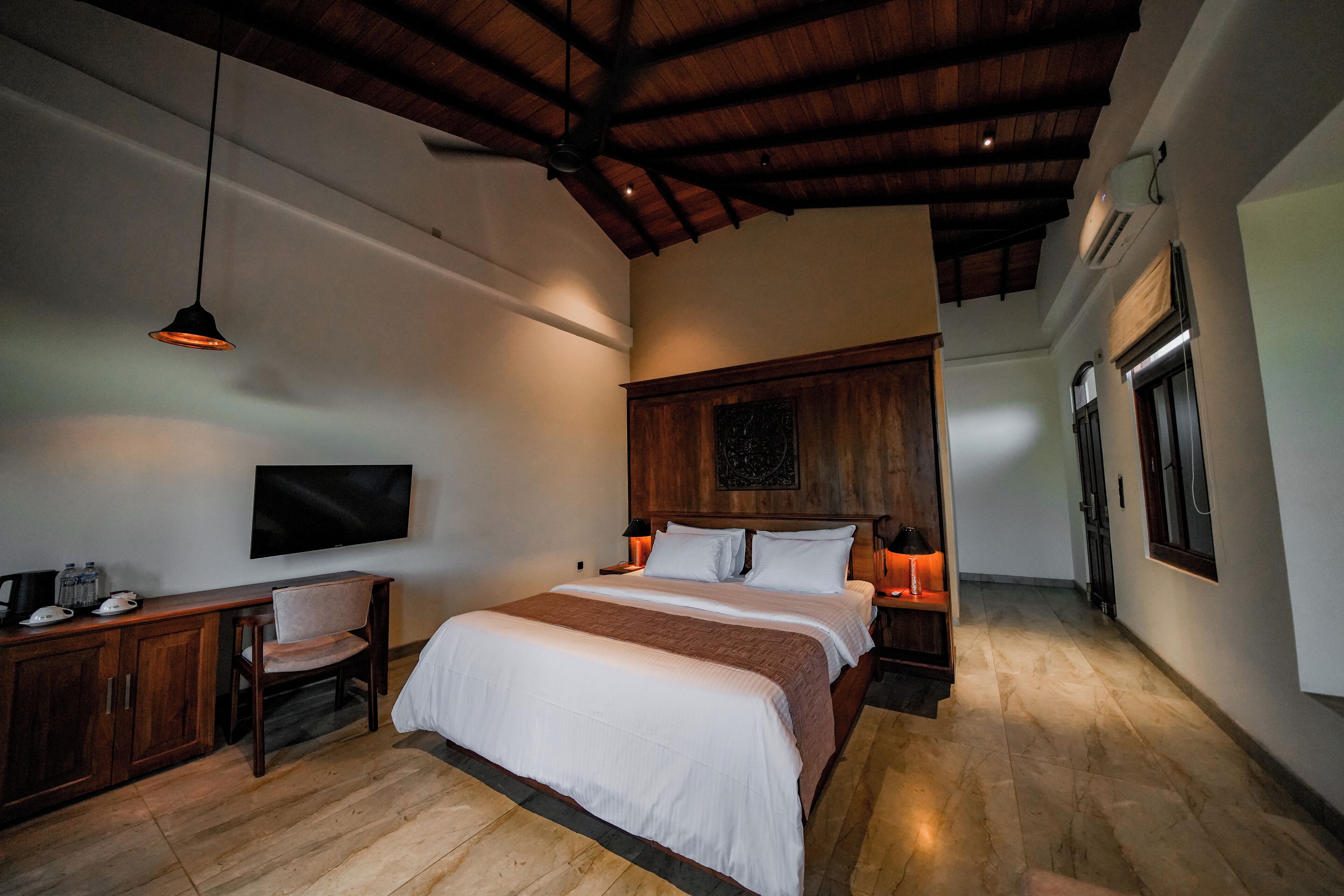 A bedroom with a wooden ceiling, a bed with white linens, a desk with a chair, and a television on the wall.