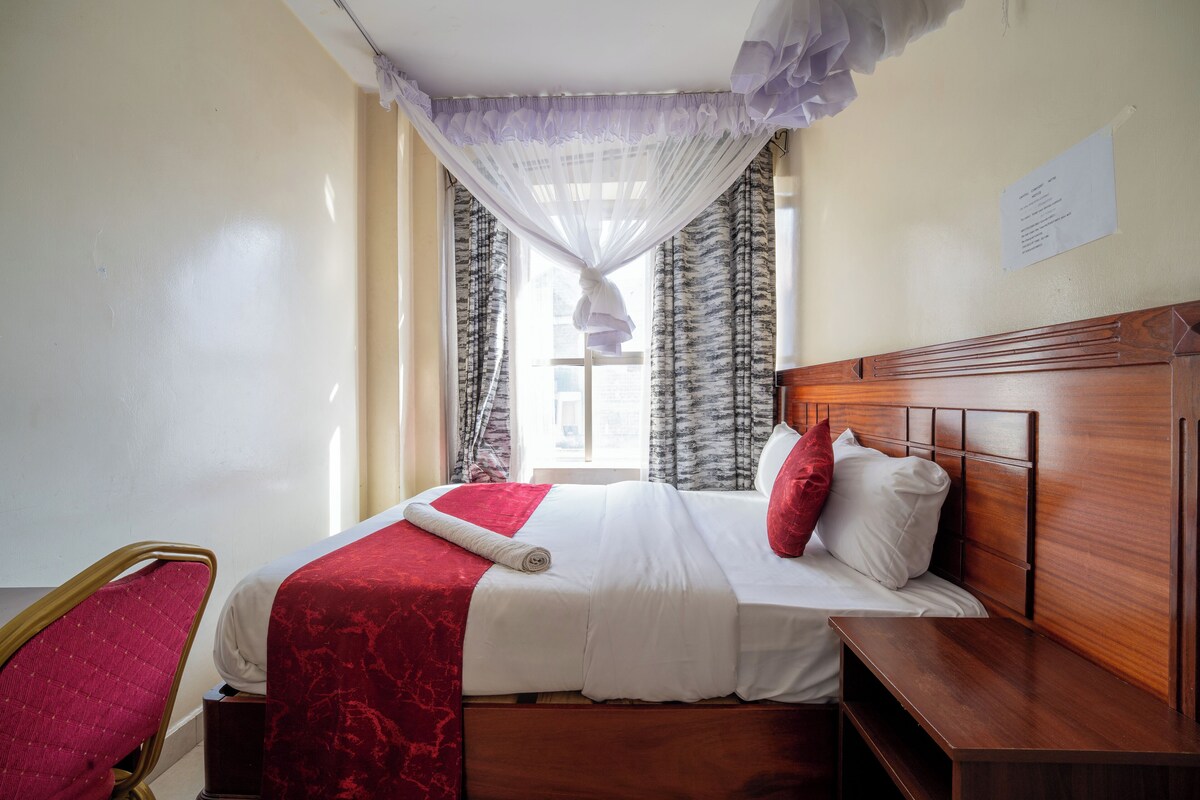 A hotel room with a wooden headboard, a bed with white and red bedding, a red chair, and a window with sheer curtains.
