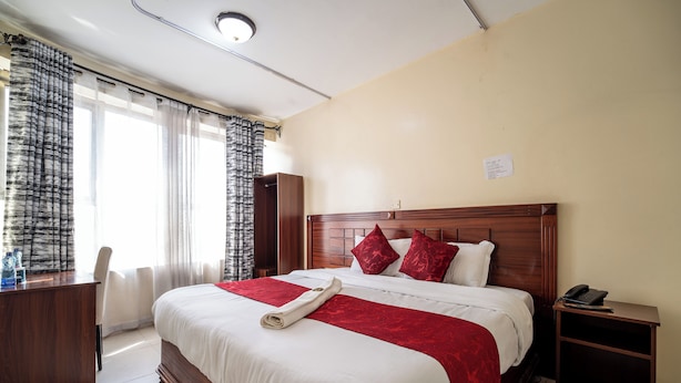 A neatly made bed with a red and white bedspread, a wooden headboard, and a bedside table with a phone. - Capital Comfort Hotel (Nairobi)