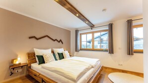 Een slaapkamer met een houten bed, een nachtkastje met een lamp, een groot raam met gordijnen en uitzicht op een gebouw.