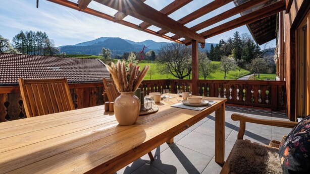 Una mesa de madera con sillas, un florero con plantas secas y vistas a un paisaje montañoso.