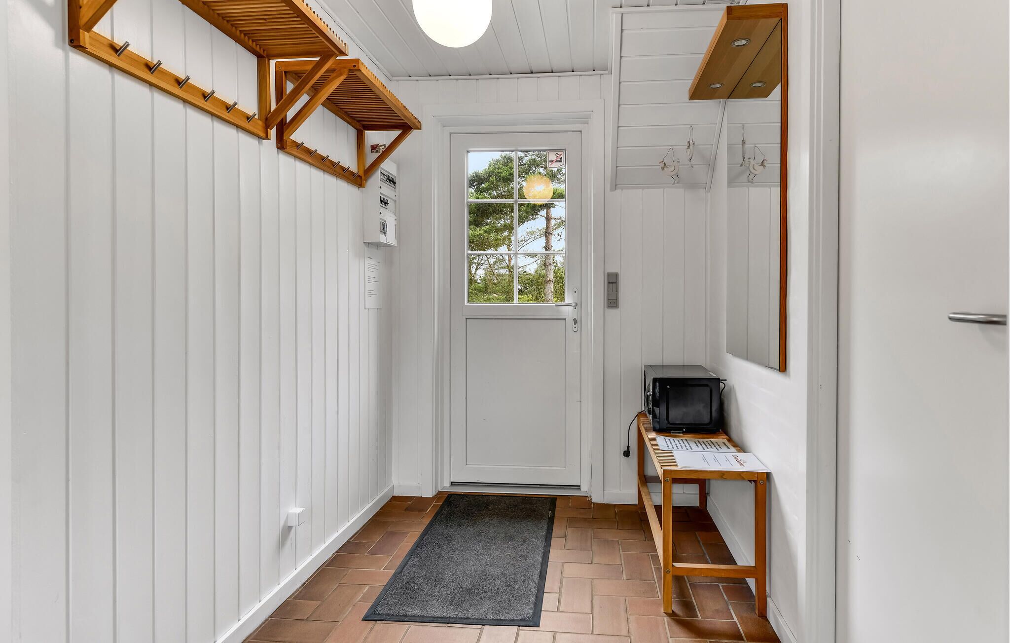Un couloir aux murs blancs, une porte en bois, une fenêtre, un paillasson noir et une table en bois sur laquelle se trouve un ordinateur.