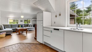 A modern kitchen with white cabinetry, a large window with a view of greenery, and a dining area with a gray sofa and wooden table. - Cozy home in Blåvand with sauna (Blåvand)