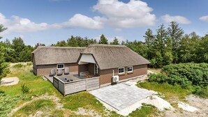 A rustic cabin with a thatched roof, a deck with lounge chairs, and a small outdoor kitchen. - Cozy home in Blåvand with sauna (Blåvand)