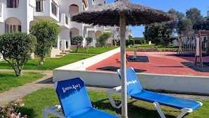 Un hotel con edificios blancos, un camino rojo y dos sillones azules con sombrilla de paja.