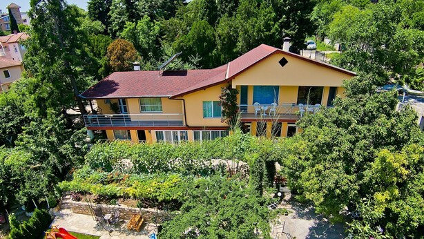 A two-story house with a red roof, surrounded by lush greenery and a small playground.
