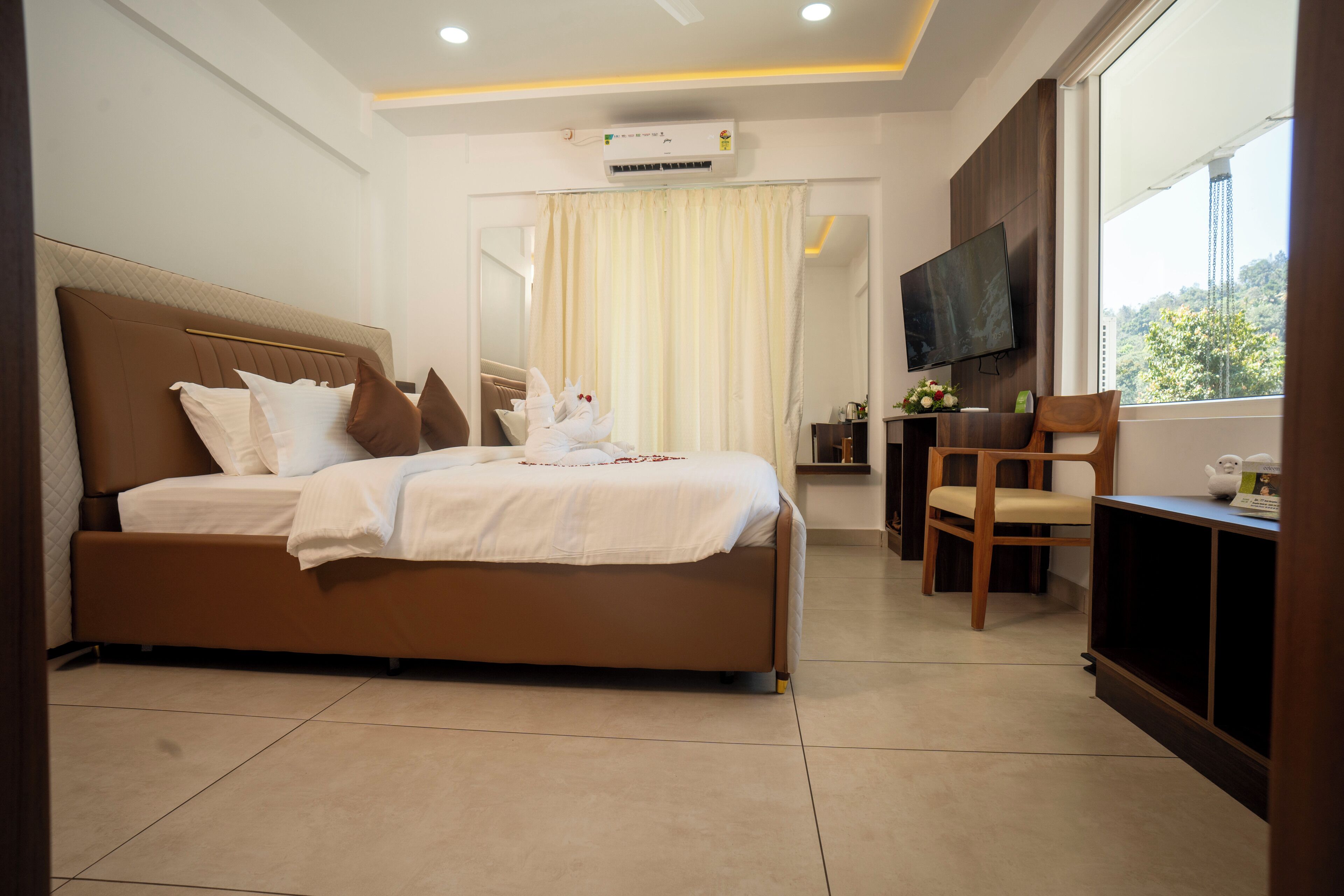 A modern hotel room with a large bed, a desk, a chair, a TV, and a window with a view of greenery.