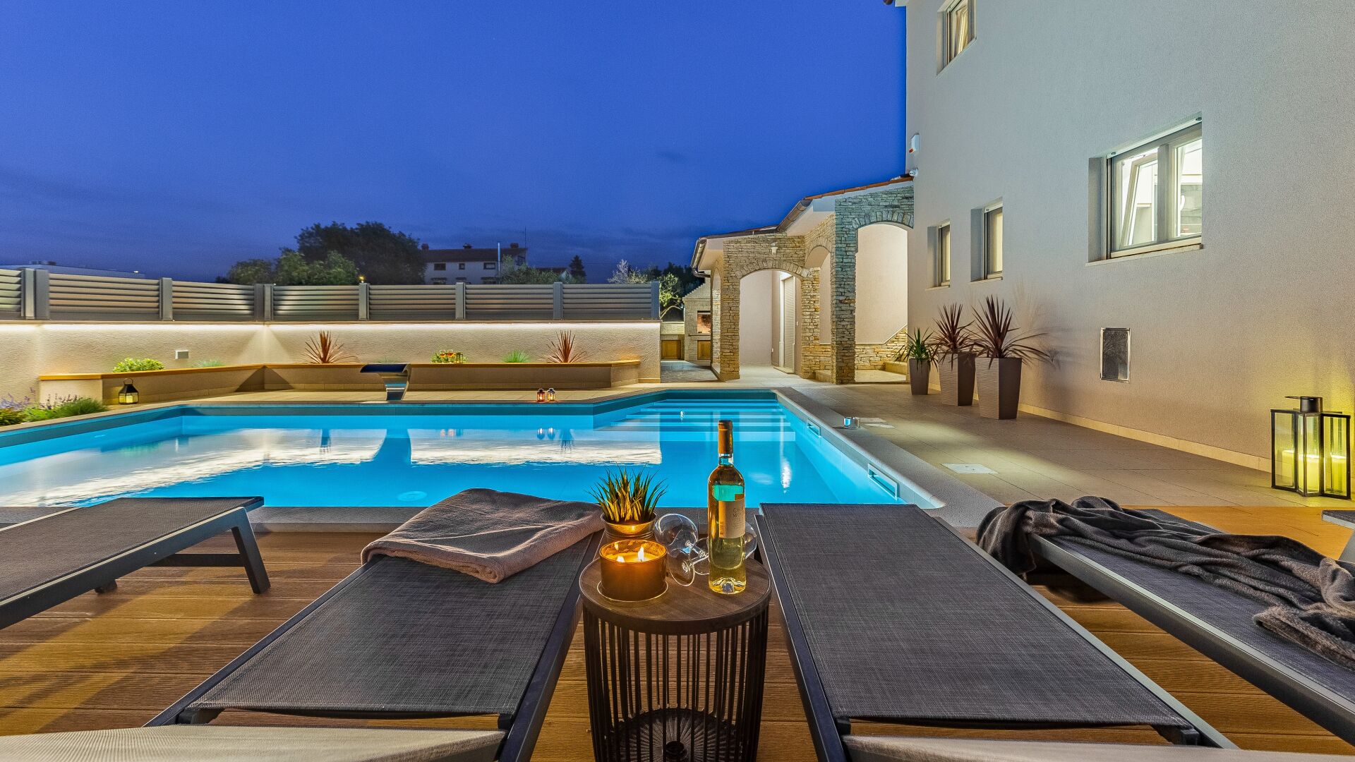 A poolside area with lounge chairs, a table, and a bottle of wine.