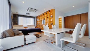 A modern living room with a sofa, ottoman, and a dining table with chairs. There is a built-in bookshelf and a large window with blinds. - Fierement inn (Sapporo)
