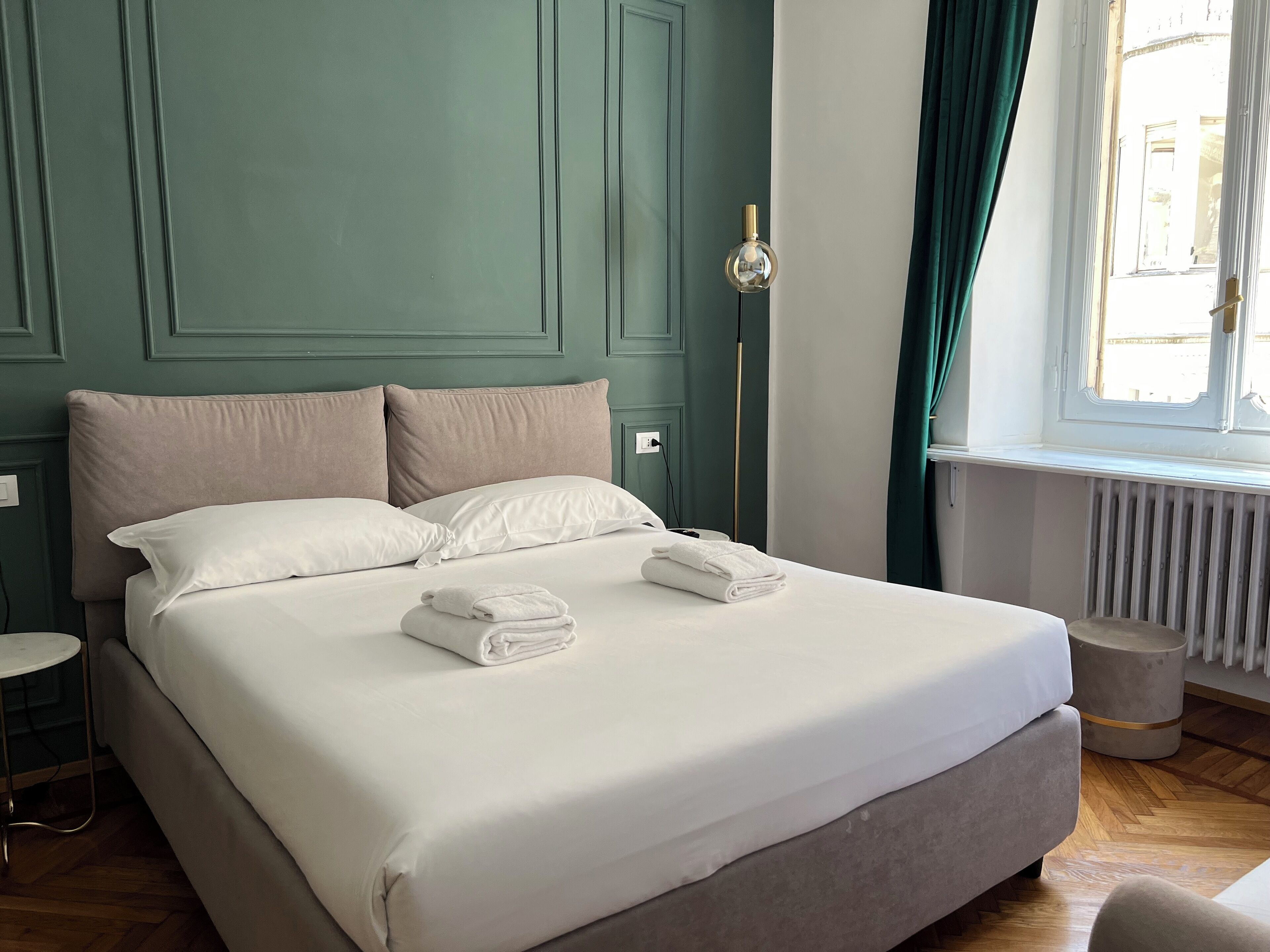 Ein ordentlich bezogenes Bett mit weißen Leinentüchern und zwei gefalteten Handtüchern, ein Nachttisch, eine Stehlampe und ein Fenster mit Vorhängen.