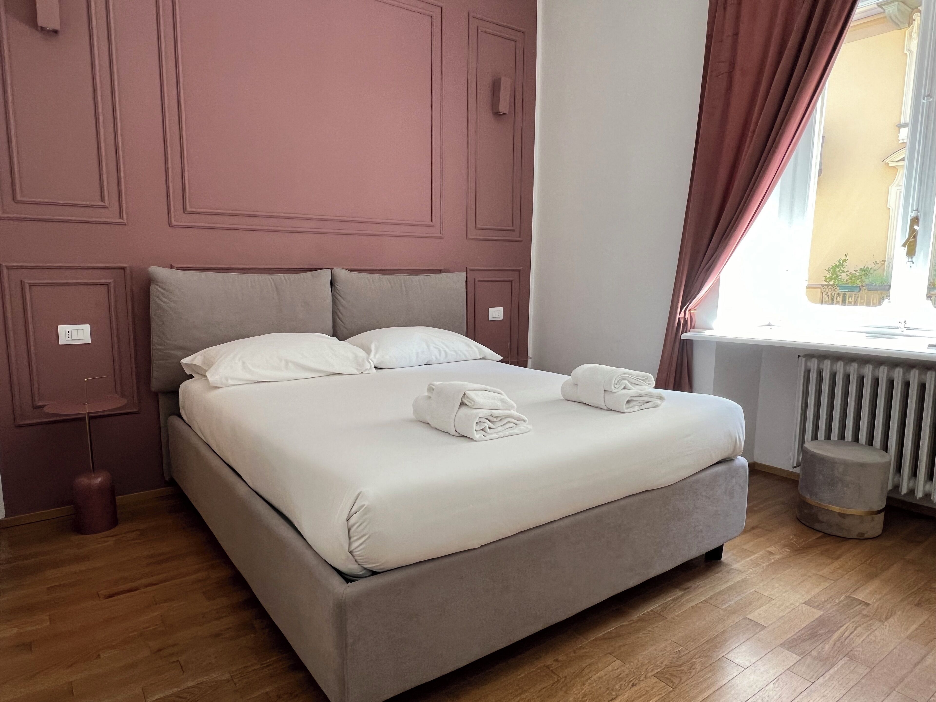 Un lit bien fait, avec des oreillers et des serviettes, un plancher en bois, un radiateur et une fenêtre avec des rideaux.