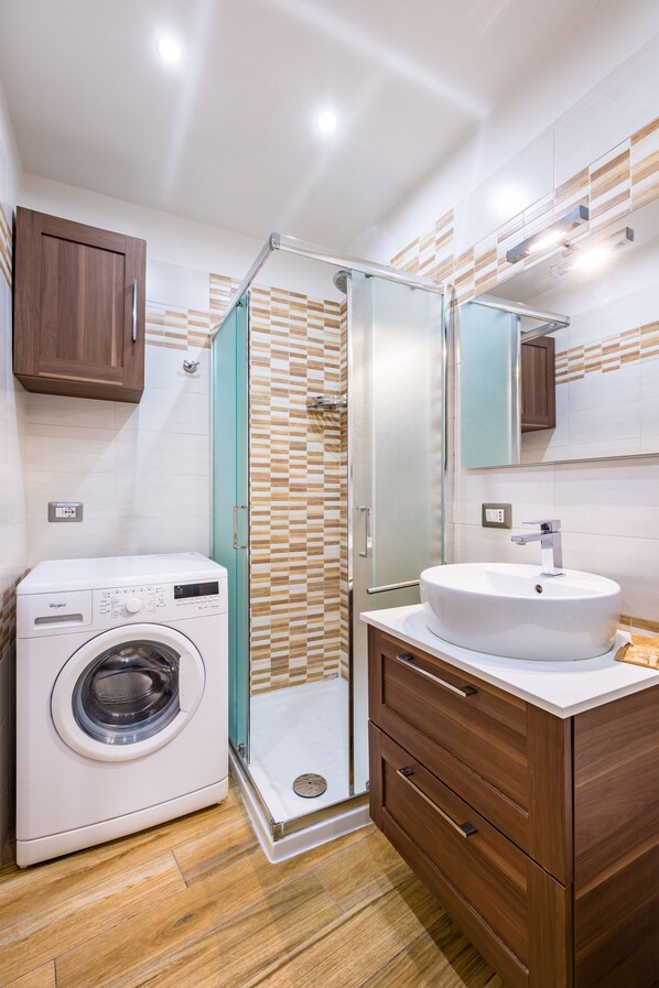 A modern bathroom with a glass shower enclosure, a built-in washing machine, and wooden cabinetry. - Cozy Escape by Napoliapartments (Naples)