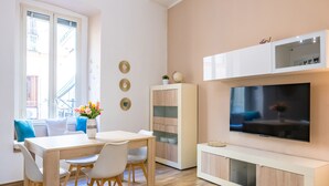 A modern living room with a wooden floor, a dining table with chairs, a TV mounted on the wall, and a ceiling light fixture. - Cozy Escape by Napoliapartments (Naples)