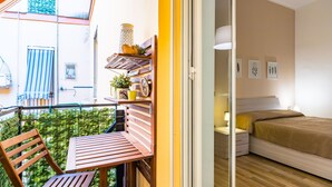 A balcony with a wooden table and chairs, a small plant, and a yellow pot. Inside, there is a bed with a brown bedspread, a nightstand, and wall art. - Cozy Escape by Napoliapartments (Naples)