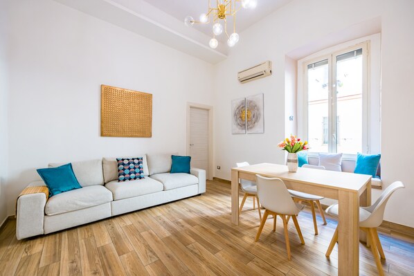 A modern living room with a sofa, dining table, and chairs. There is a window with a view, a ceiling light, and a wall-mounted air conditioner. - Cozy Escape by Napoliapartments (Naples)