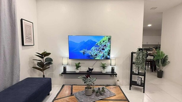 A modern living room with a flat-screen TV, a blue sofa, a coffee table with a game, and a kitchen in the background.