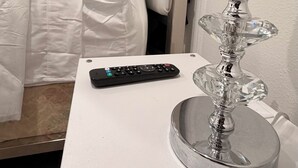 Una cama bien tendida con ropa blanca, un control remoto y una lámpara de cristal decorativa sobre una mesita de noche blanca.
