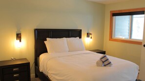 A neatly made bed with white linens and pillows, two bedside tables with lamps, and a window with blinds. - Fernie - Timberline Lodges Spruce #236 (Fernie)