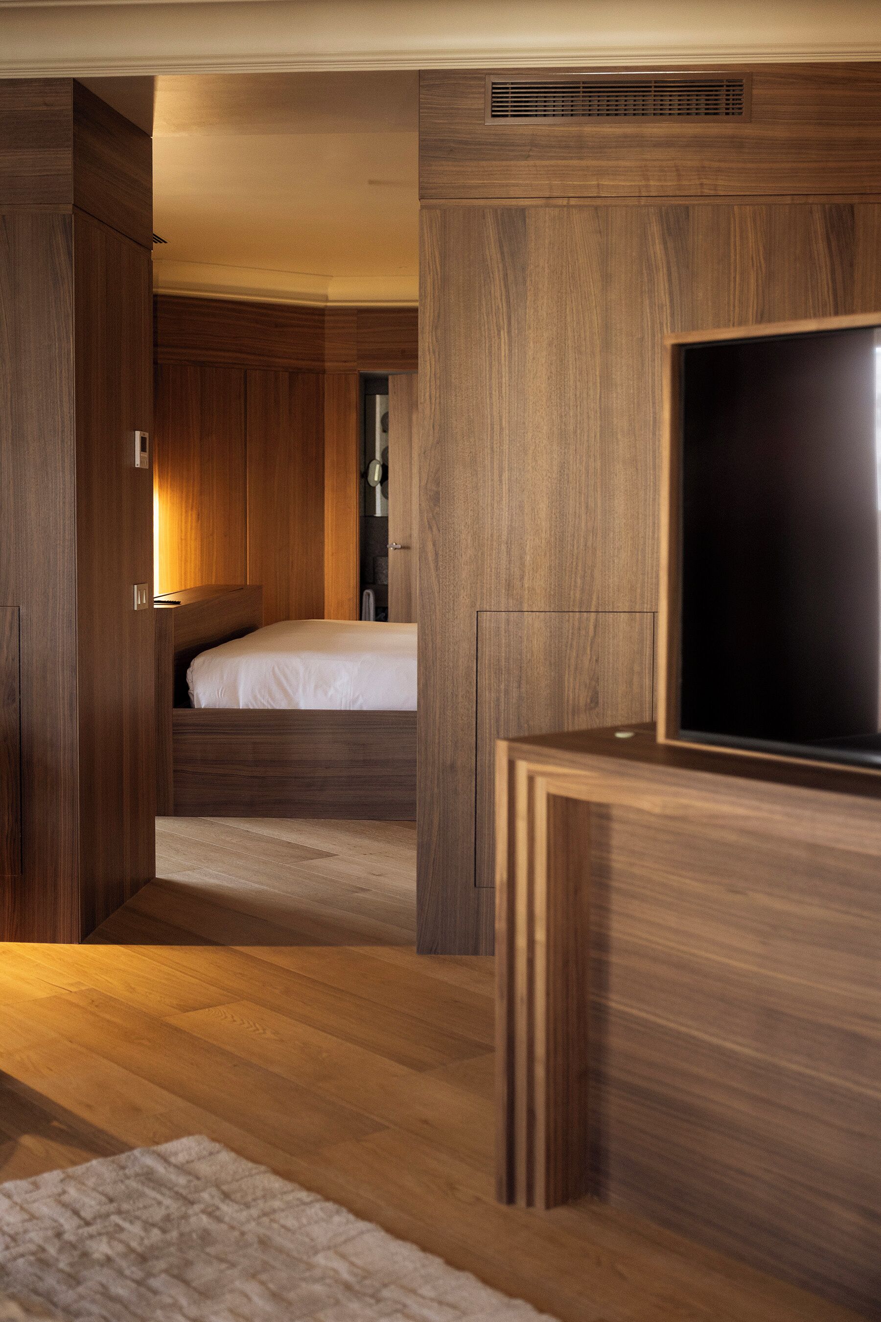 A modern hotel room with a wooden floor, a bed, a television, and built-in wooden cabinets.