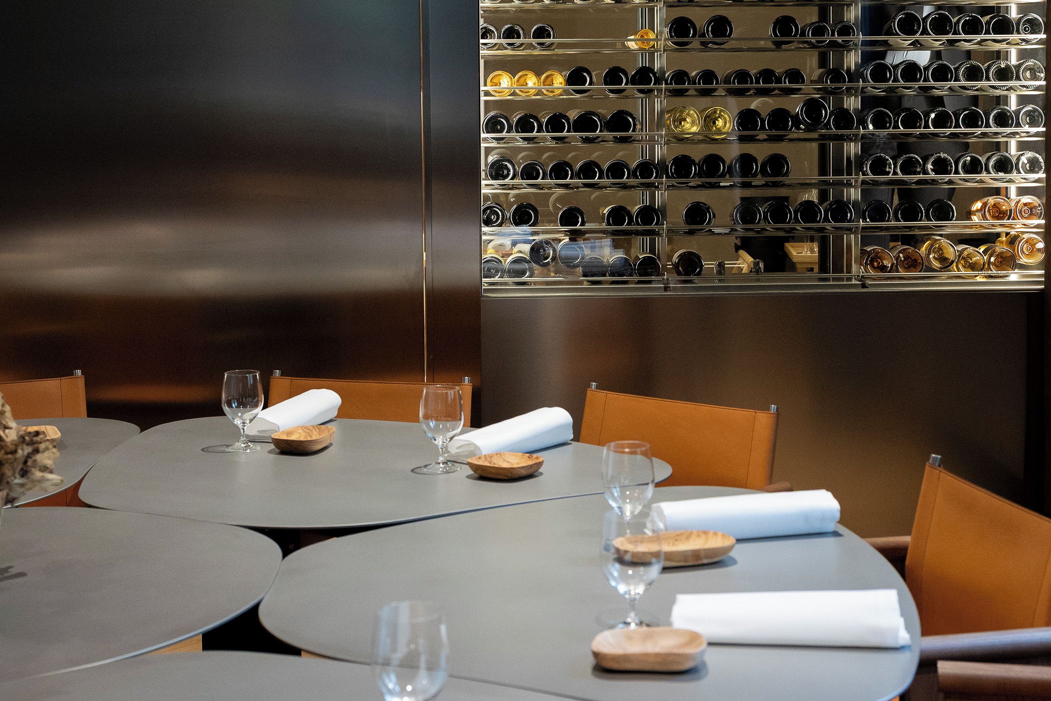 L’intérieur d’un restaurant avec des tables rondes, des verres à vin et un présentoir à bouteilles en arrière-plan.