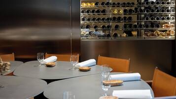 Interior de restaurante con mesas redondas, copas de vino y una estantería para botellas en el fondo.