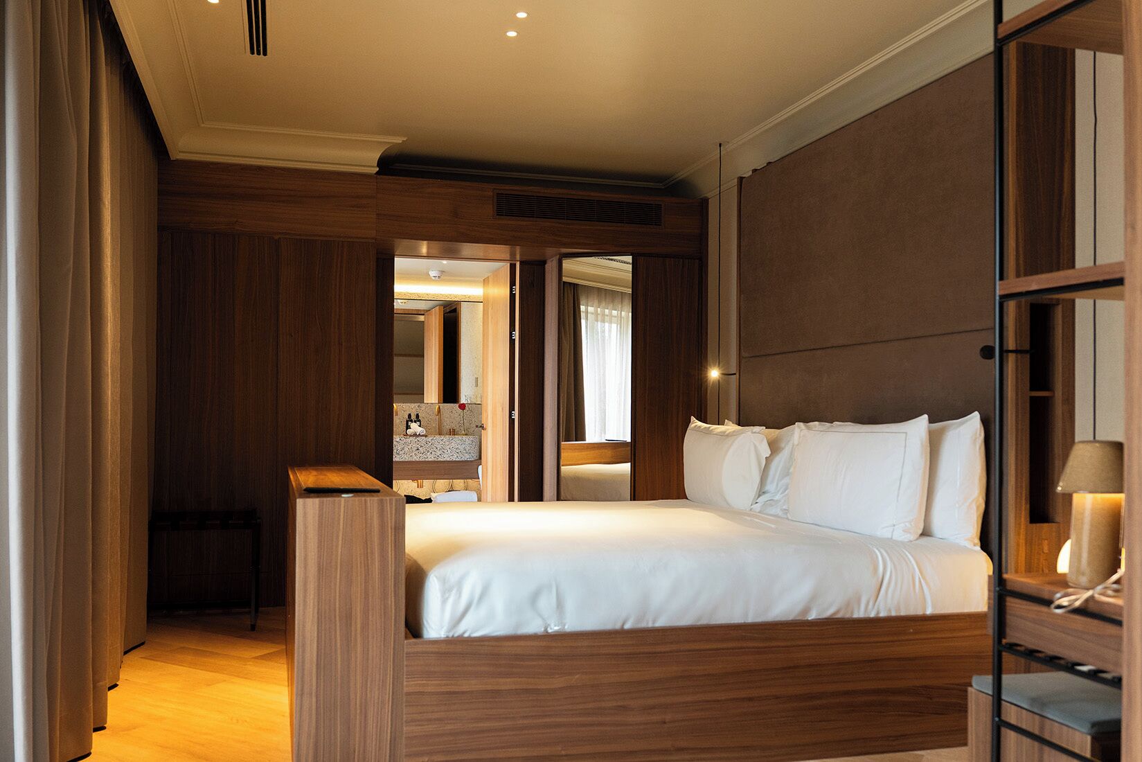 Habitación de hotel con cama de madera, mesita de noche con lámpara, espejo y vistas al baño.