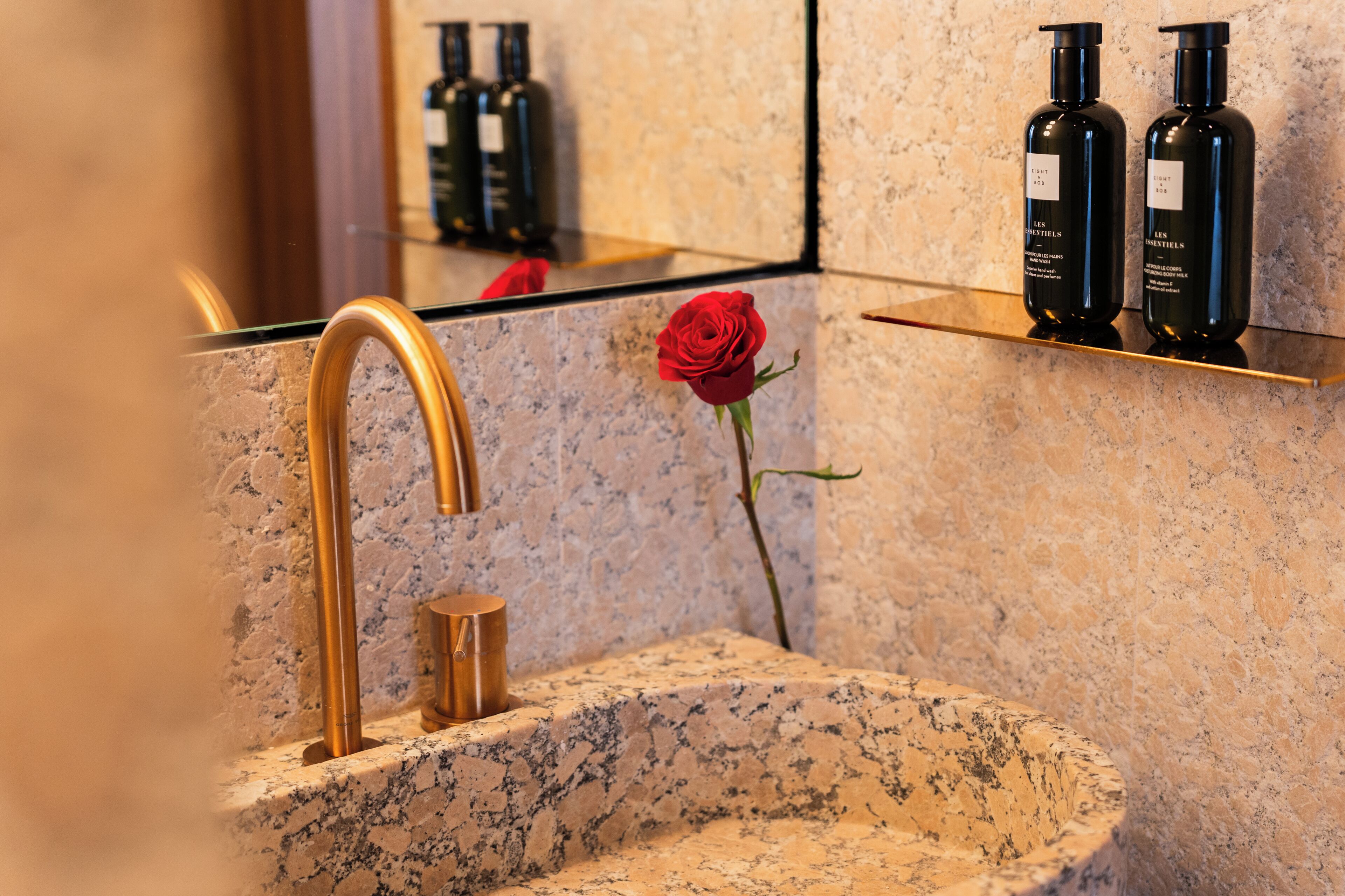 A bathroom with a granite sink, a gold faucet, a red rose, and two black bottles on a shelf.
