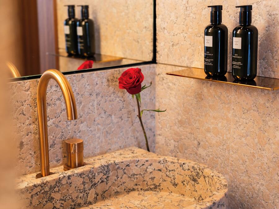 A bathroom with a granite sink, a gold faucet, a red rose, and two black bottles on a shelf.