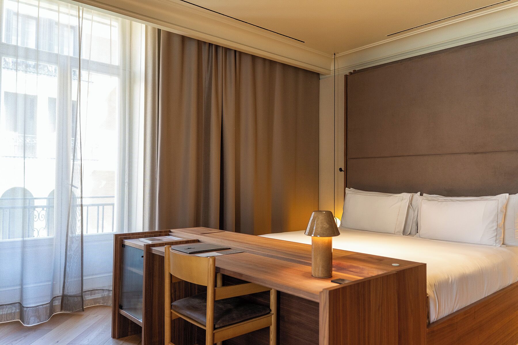A hotel room with a bed, a desk, a chair, a lamp, and a view of the outside through a window.