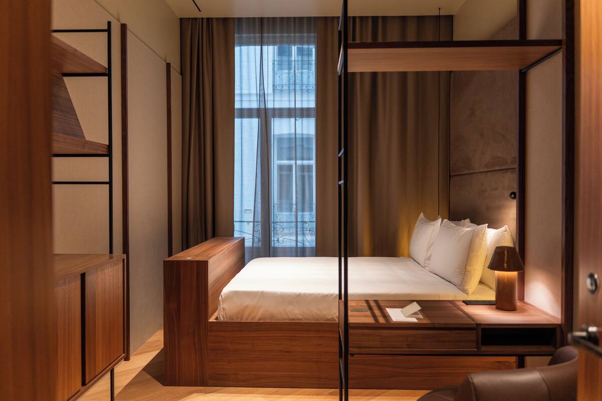 Een moderne slaapkamer met een houten bedframe, een nachtkastje, een lamp en uitzicht op de stad door grote ramen.
