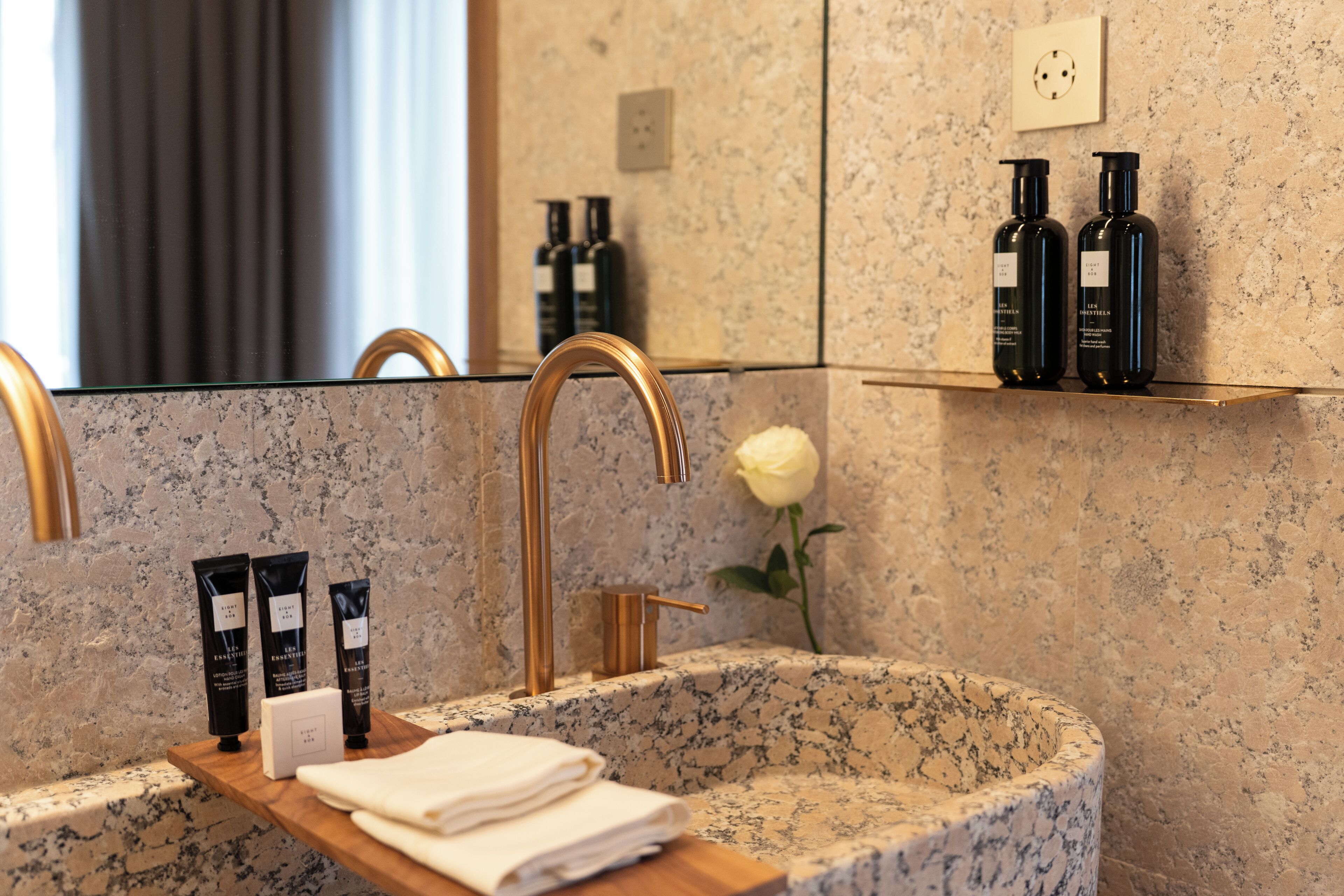 A bathroom with a granite sink, gold faucet, and a mirror reflecting the room. There are two black bottles on a shelf above the sink and a white rose on the counter.
