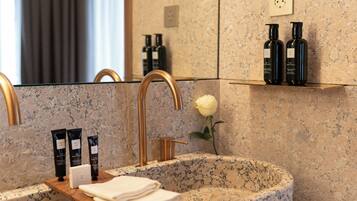 A bathroom with a granite sink, gold faucet, and a mirror reflecting the room. There are two black bottles on a shelf above the sink and a white rose on the counter.