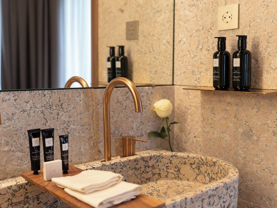 A bathroom with a granite sink, gold faucet, and a mirror reflecting the room. There are two black bottles on a shelf above the sink and a white rose on the counter.