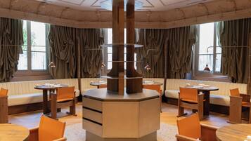Ein Restaurantbereich in einem Hotel mit einer zentralen Holzsäule, runden Tischen und gepolsterten Sitzgelegenheiten.