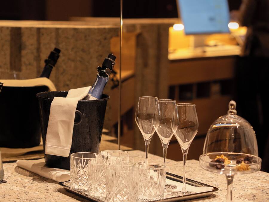 Una zona de bar con una botella de champán, copas, un ordenador portátil y una persona al fondo.