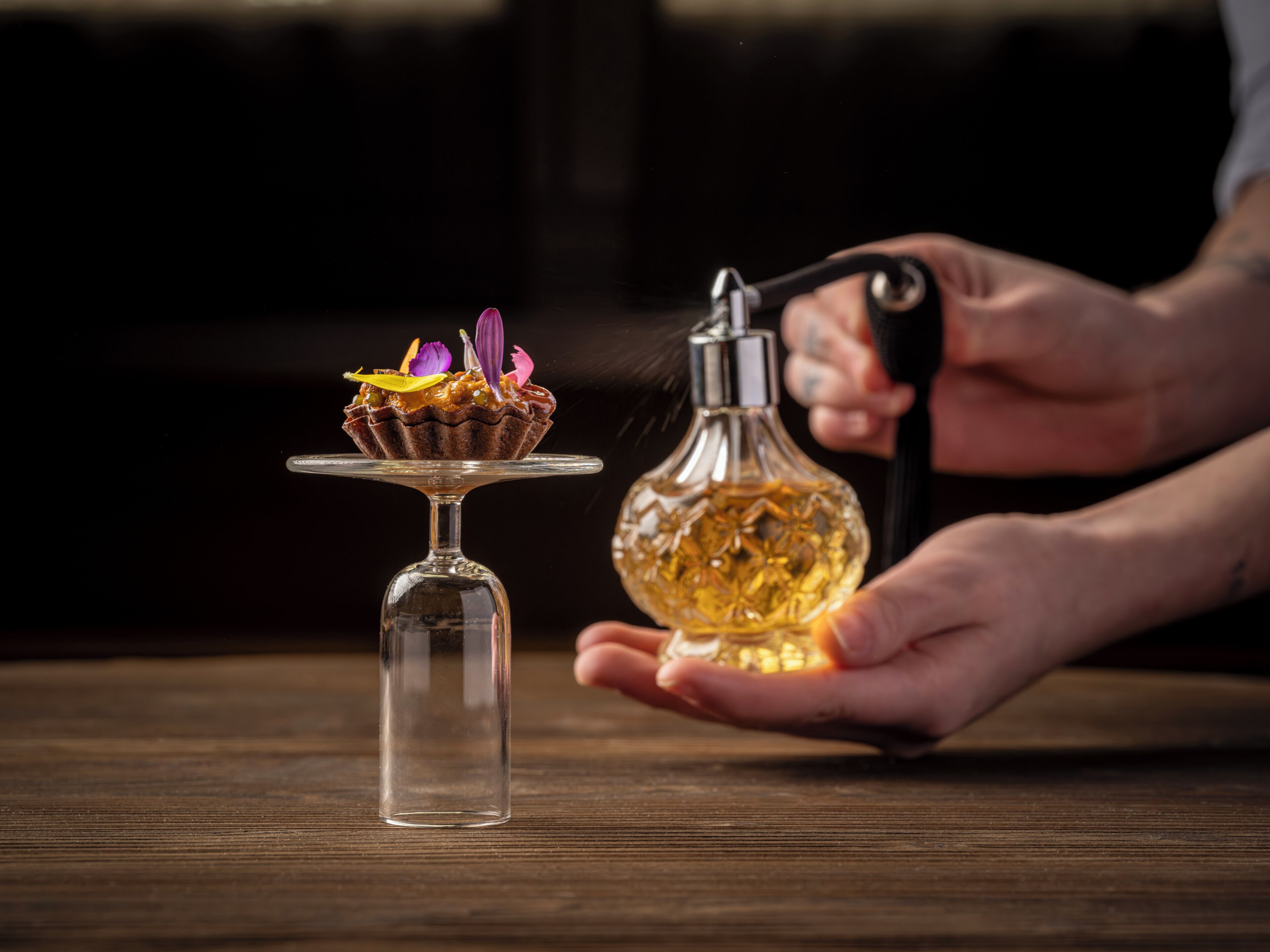 Una mano sostiene una botella de vidrio con una flor, un tallo de vidrio y un postre sobre una superficie de madera.