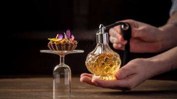 Una mano sostiene una botella de vidrio con una flor, un tallo de vidrio y un postre sobre una superficie de madera.