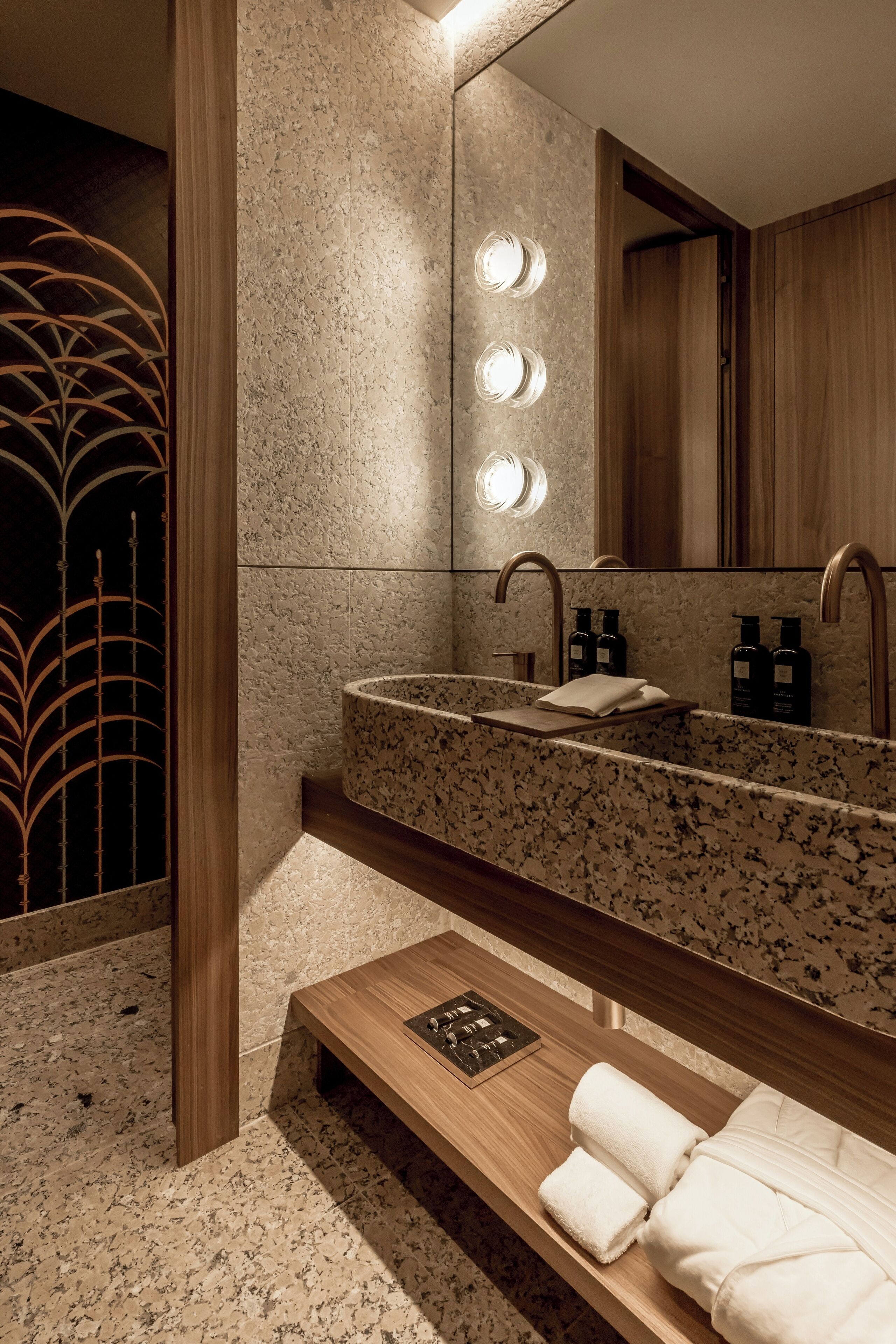 A modern bathroom with a granite countertop sink, a wooden vanity, a bench with towels, and a large mirror with lights.