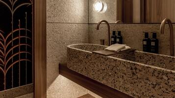 A modern bathroom with a granite countertop sink, a wooden vanity, a bench with towels, and a large mirror with lights.