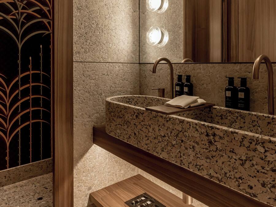 A modern bathroom with a granite countertop sink, a wooden vanity, a bench with towels, and a large mirror with lights.